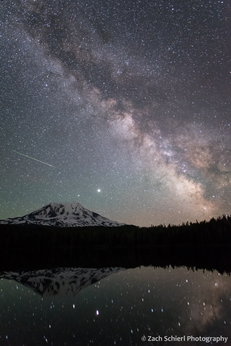 The night sky including the Milky Way and the streak of a meteor is seen over a tall mountain peak.