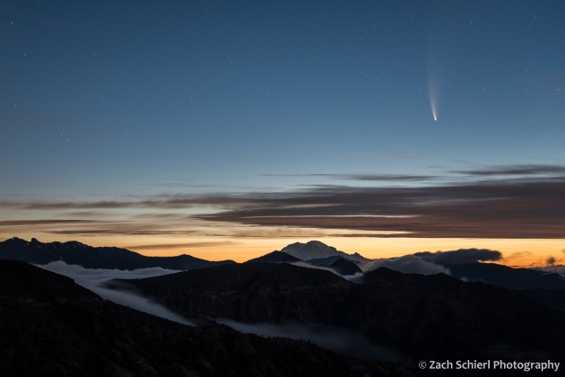 A comet and its tail appears in the pre-dawn sky with a mountain range and valley fog in the foreground