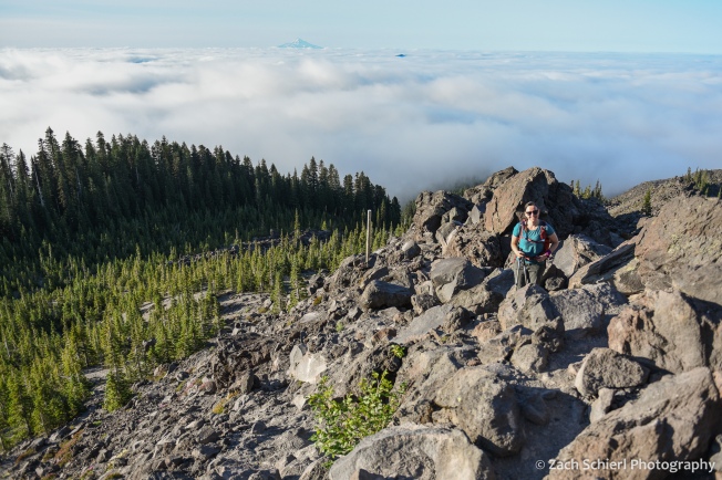 A hiker scrambles up a rocky slope with a forest and low-lying clouds in the background