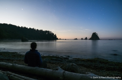 A person sits on a log along the beach watching the moon rise over the coastline.