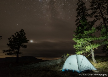 A softly lit tent at the edge of a forest is seen with the night sky partially obscured by clouds overhead.